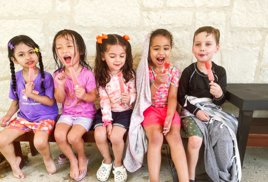 Five children on a bench, each eating a popsicle, smiling. Outdoor setting.