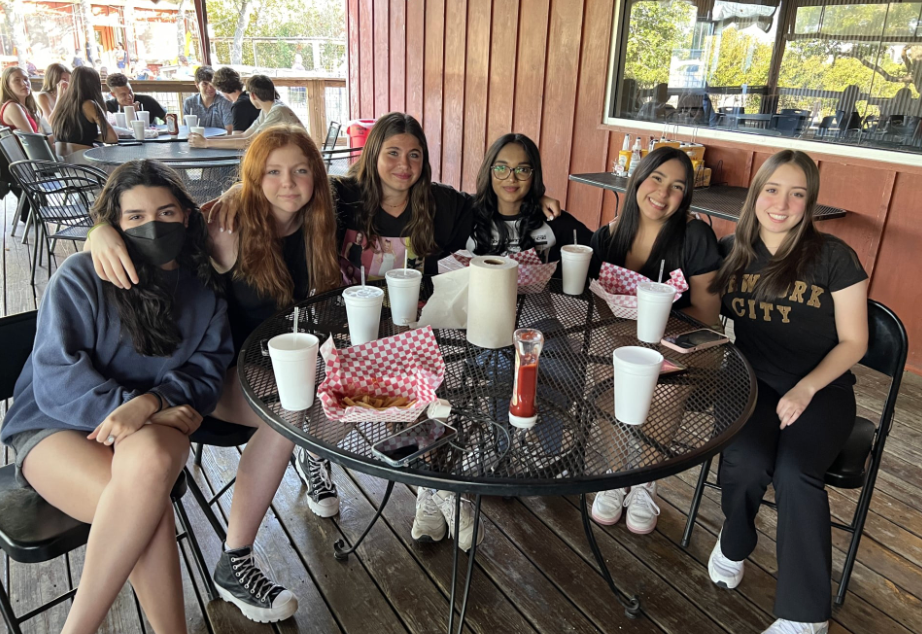 Six young people at a table on a patio. They sit, smile, and look at the camera. Food and drinks are on the table.