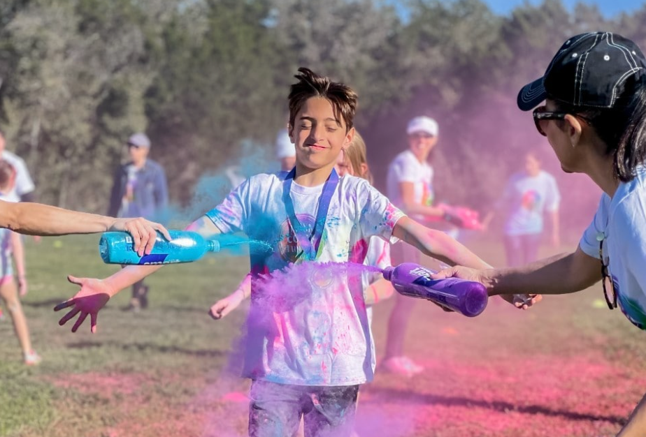 Boy covered in colorful powder during a race, others spraying him with color, outdoors.