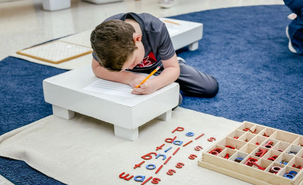 A child writes on a paper set on a low white table; wooden letter tiles are nearby on a rug.