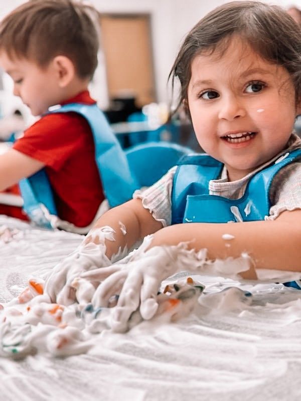 Child smiling, playing with soapy, colorful hands at a table, another child in the background.