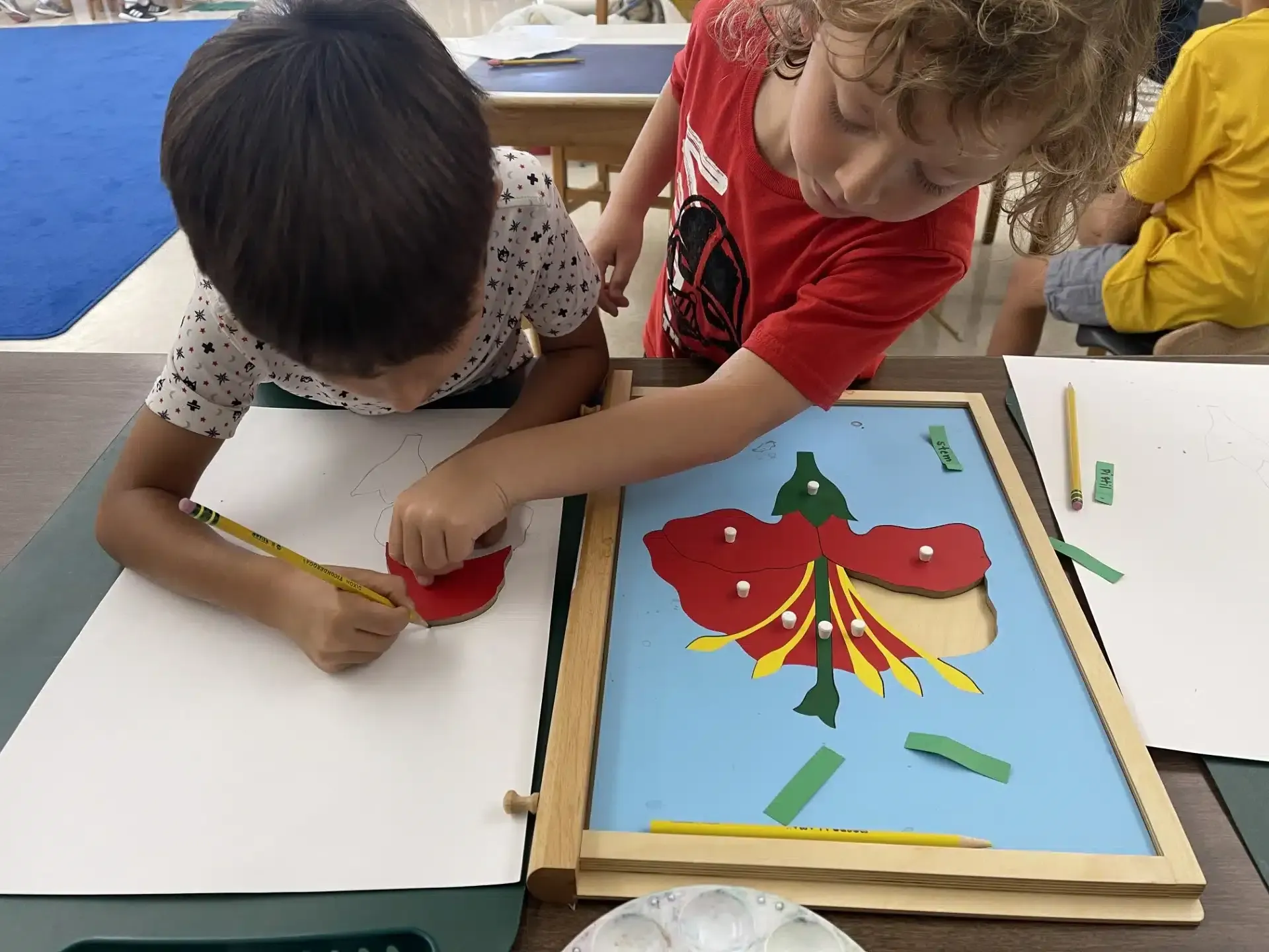 Two children drawing and working with a wooden puzzle. One draws a red shape while another points.