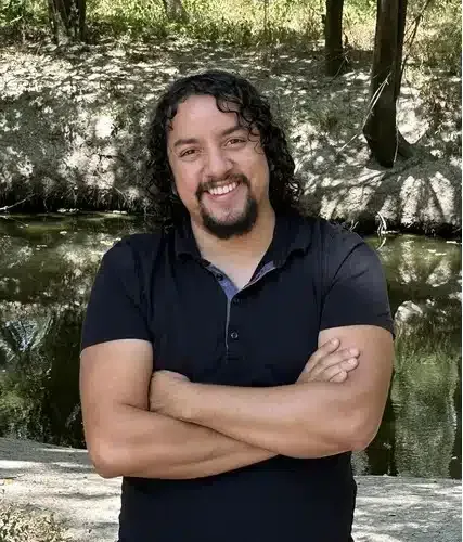 Man with long curly hair and a beard smiles, arms crossed, standing outdoors by a stream.