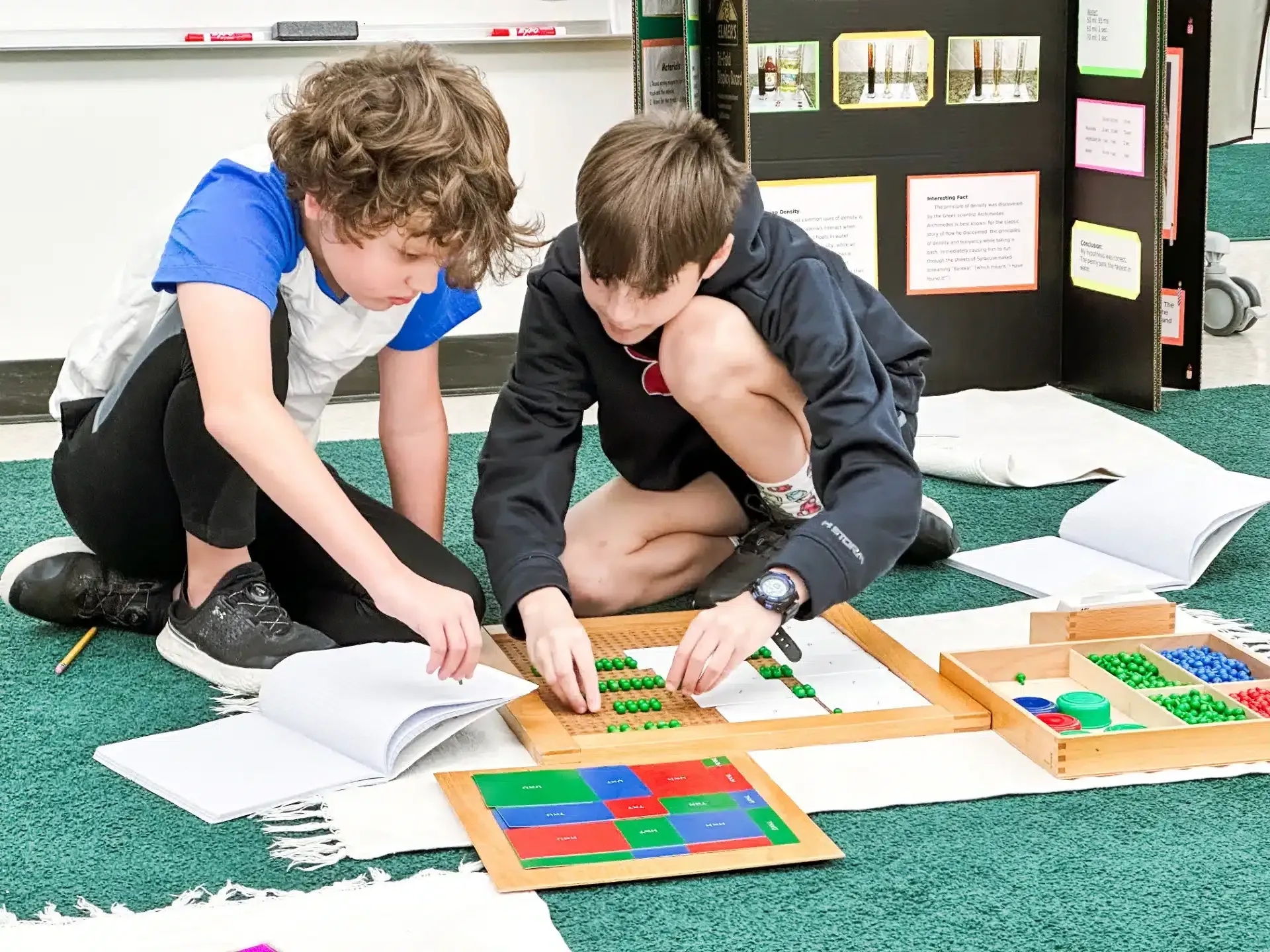 Two children work with math manipulatives on a rug, near a presentation board.