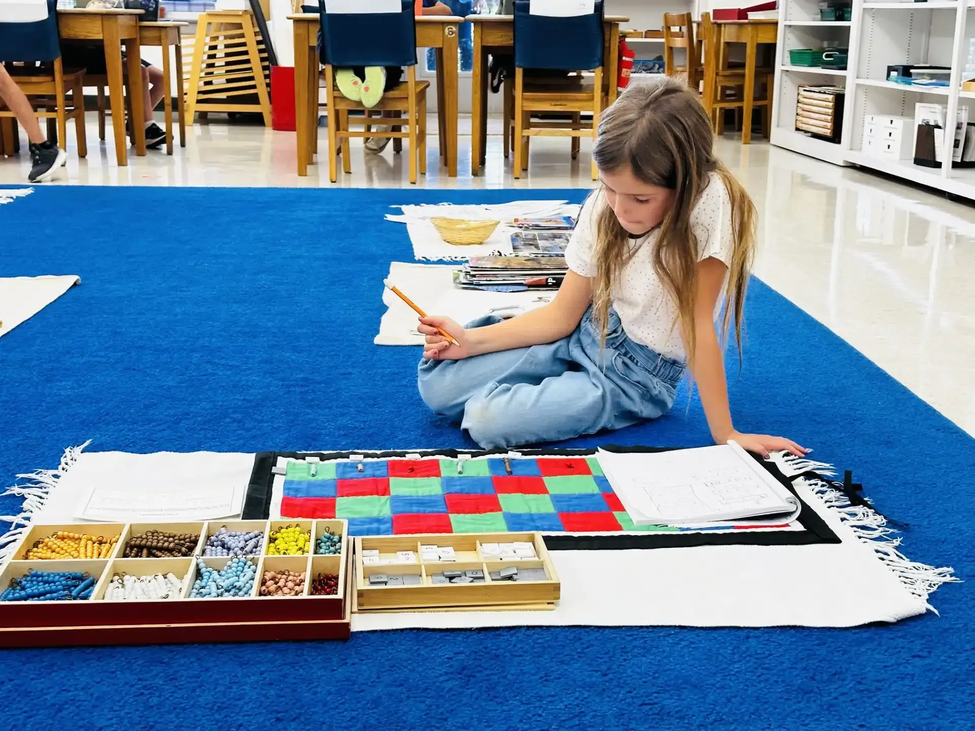 A girl kneels on a blue rug, working with educational materials: blocks, beads, and a notebook in a classroom.