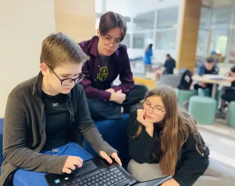 Three people looking at laptop, indoors. One points; others watch, thoughtful expressions. Blue sofa, bright background.