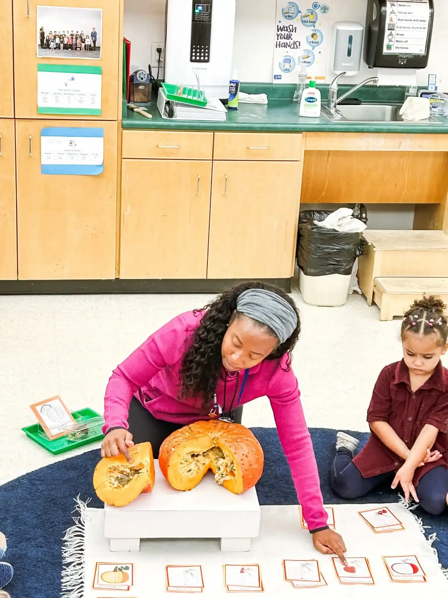 Teacher and student examining a pumpkin, cards in front of them, in a classroom setting.