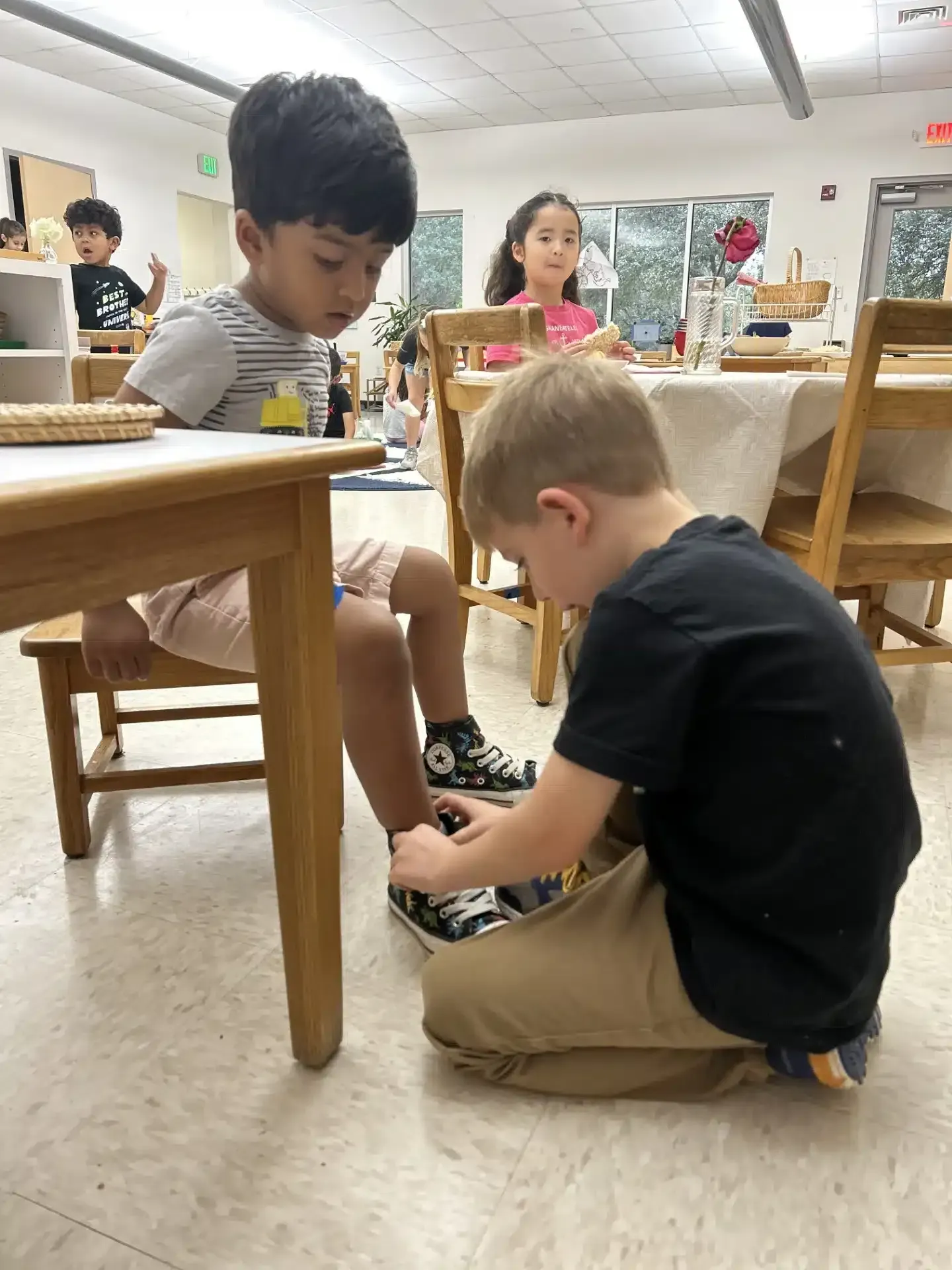 Boy ties friend's shoe in a classroom. Other children and tables in the background.