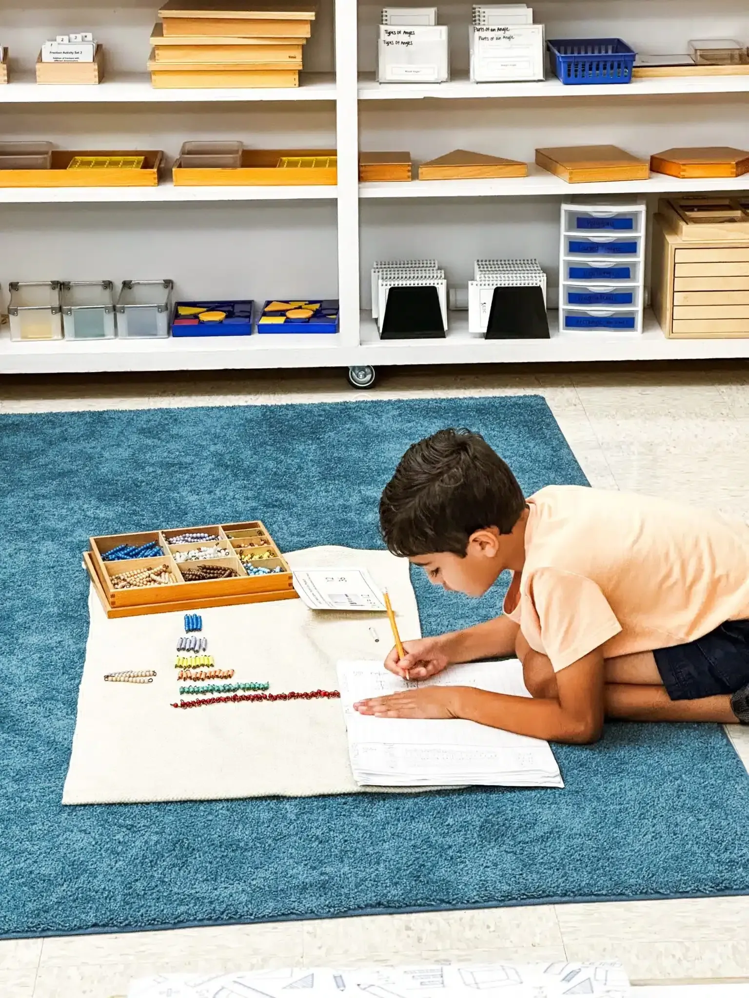 Boy writing on paper on a blue rug, a Montessori classroom shelf in the background.