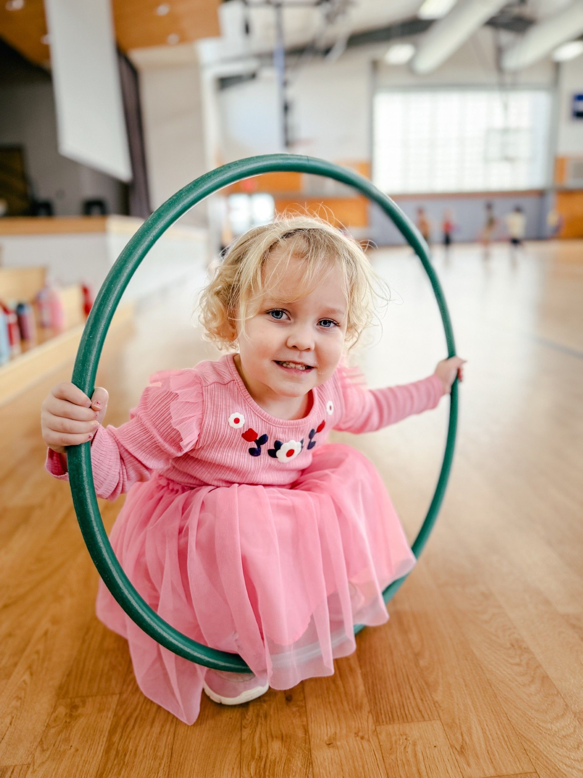 A girl in a pink dress in a gymnasium hunched over a green hula-hoop.
