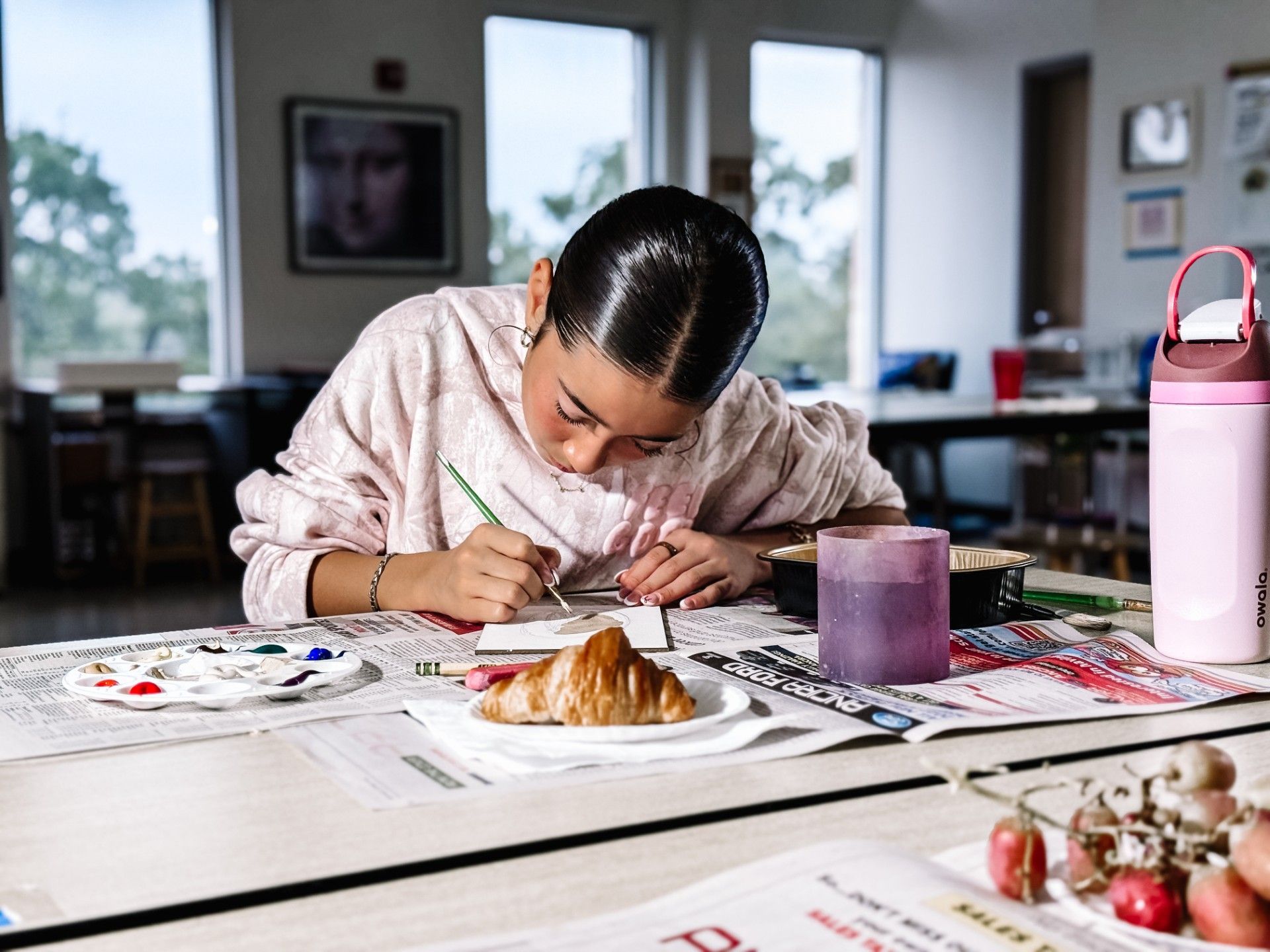 Girl in a pink sweatshirt with a tray of paint and a croissant is painting the croissant.