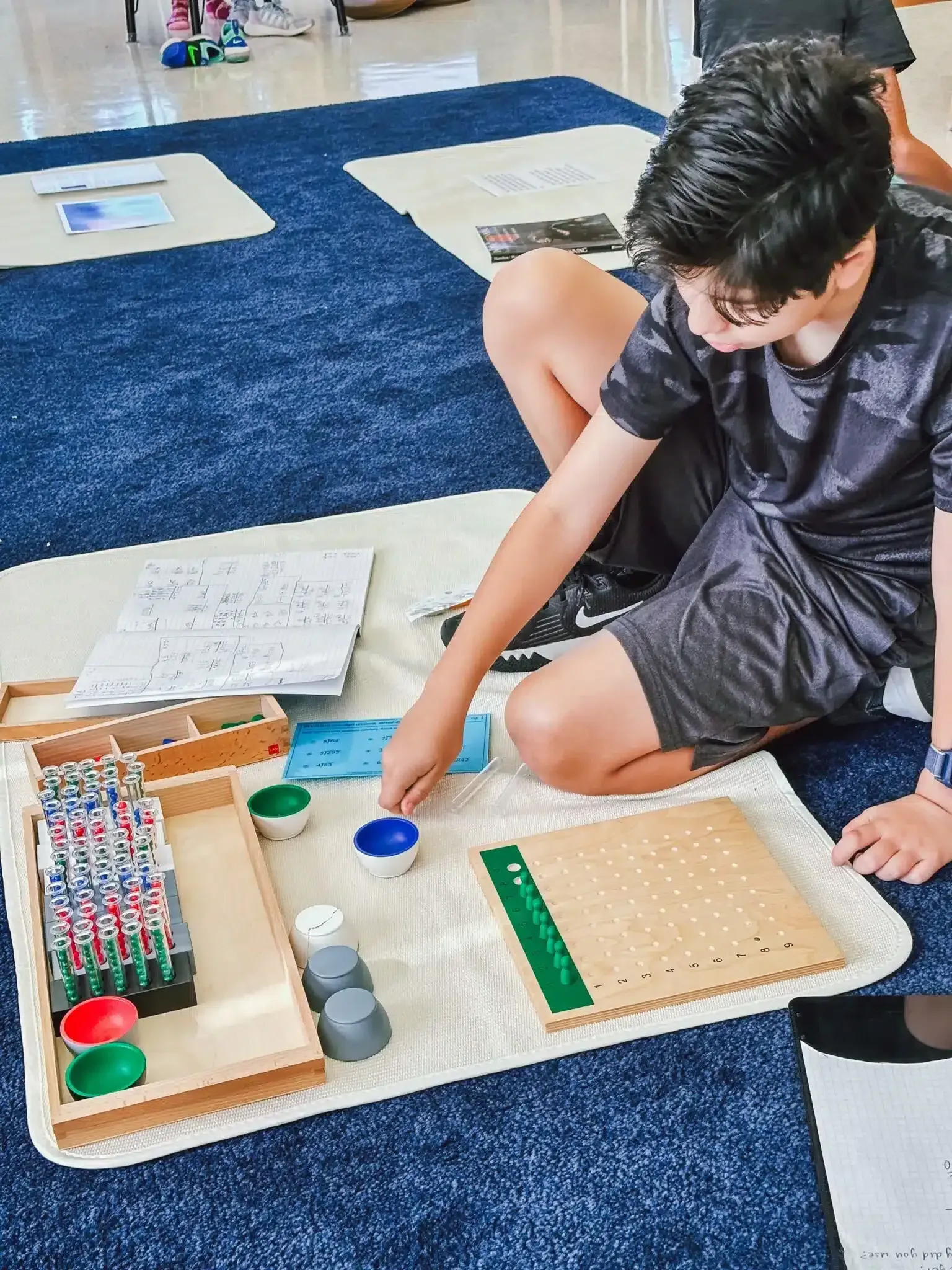 Boy using Montessori math materials on a rug; counting with colored beads and wooden boards.