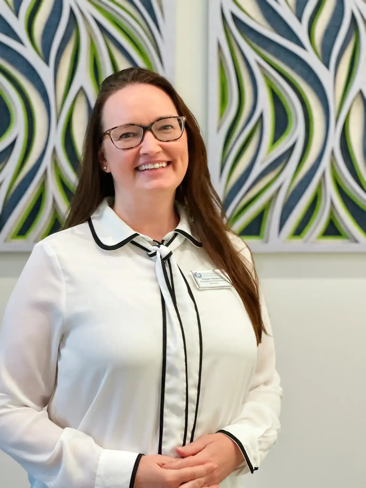 Woman with glasses smiling, wearing a white blouse with black trim, standing in front of wall art.