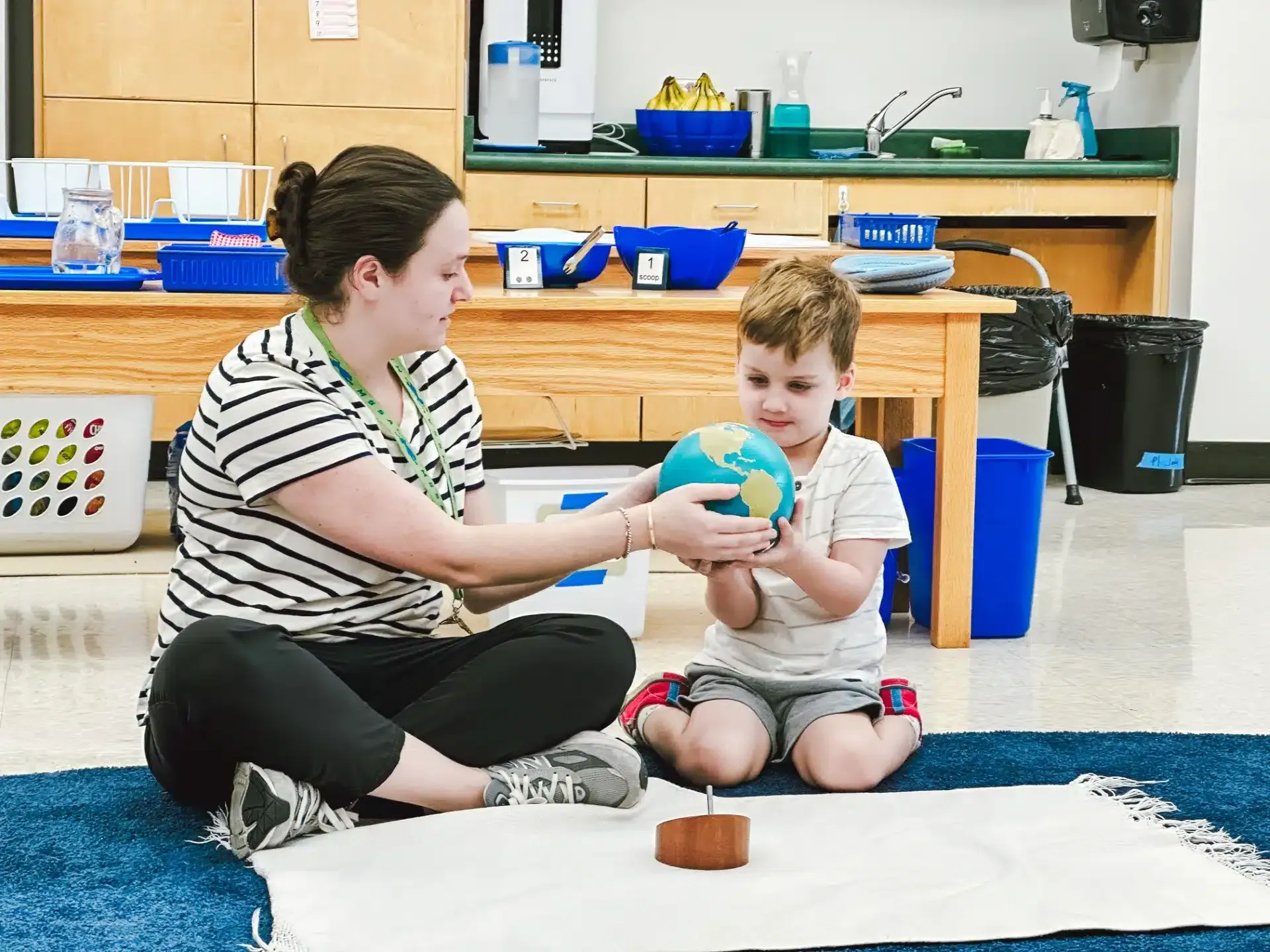 Teacher and child examining a globe together, seated on the floor in a classroom.