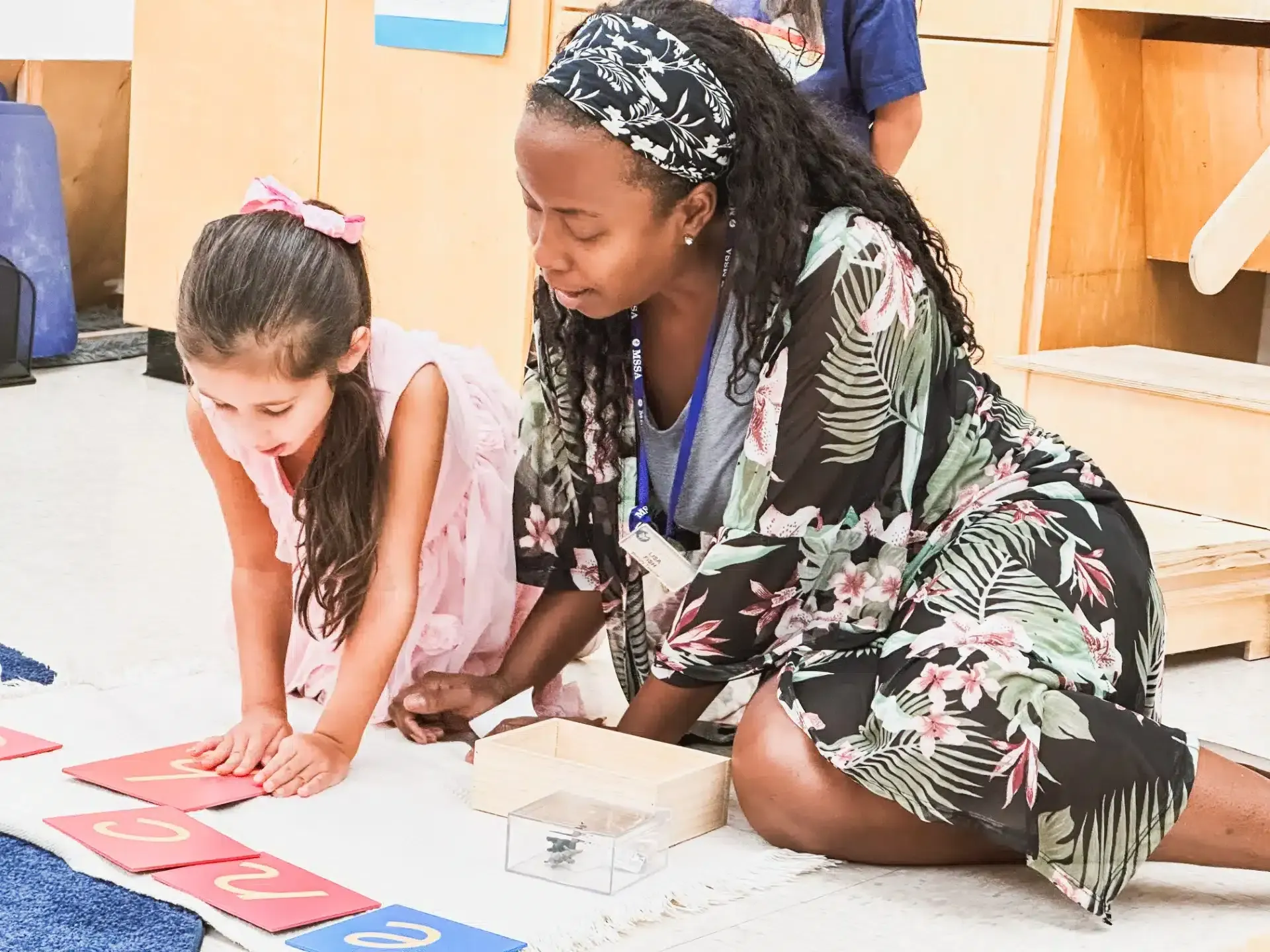 Woman assisting child with number cards on a rug in a classroom.