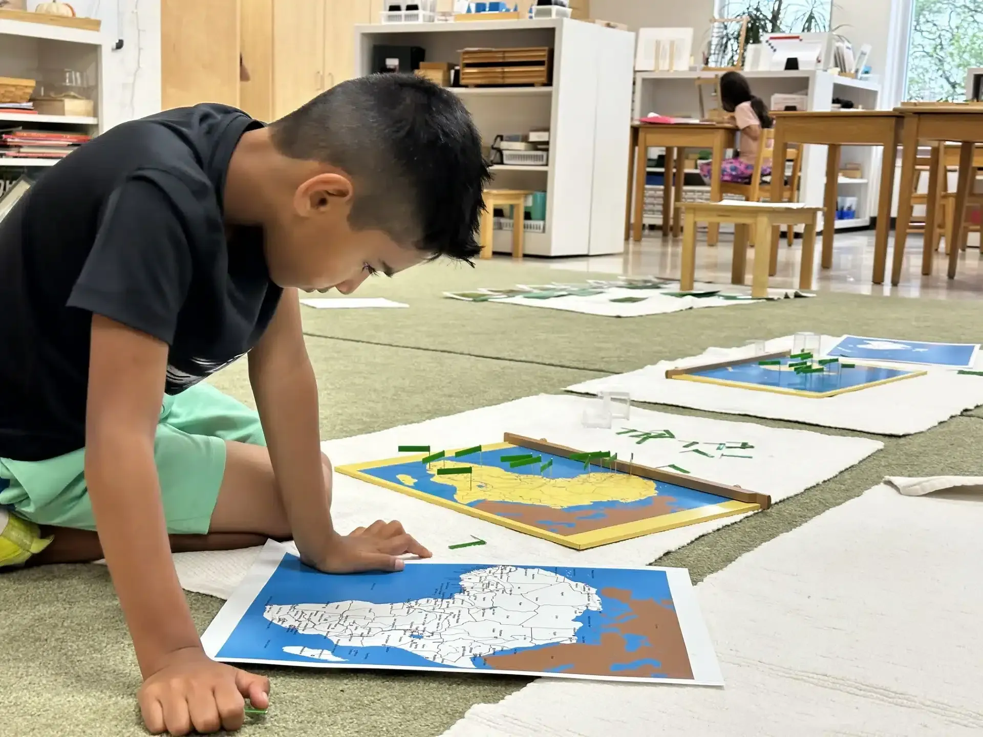 Boy on floor with map puzzles, focusing intently. Classroom setting with other students visible.
