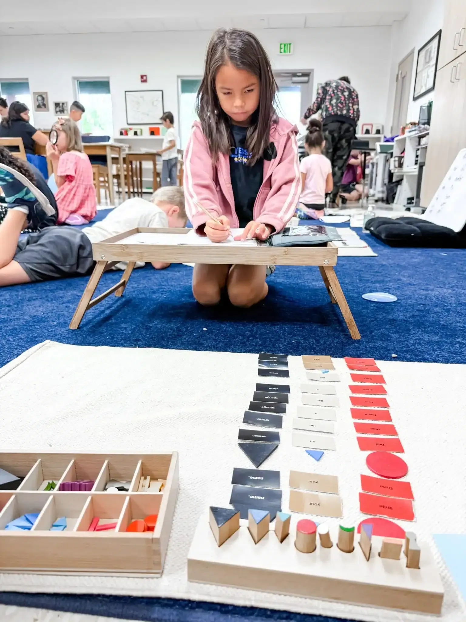 Child kneeling at low desk, drawing. Colorful shapes on mats in classroom. Other children in background.