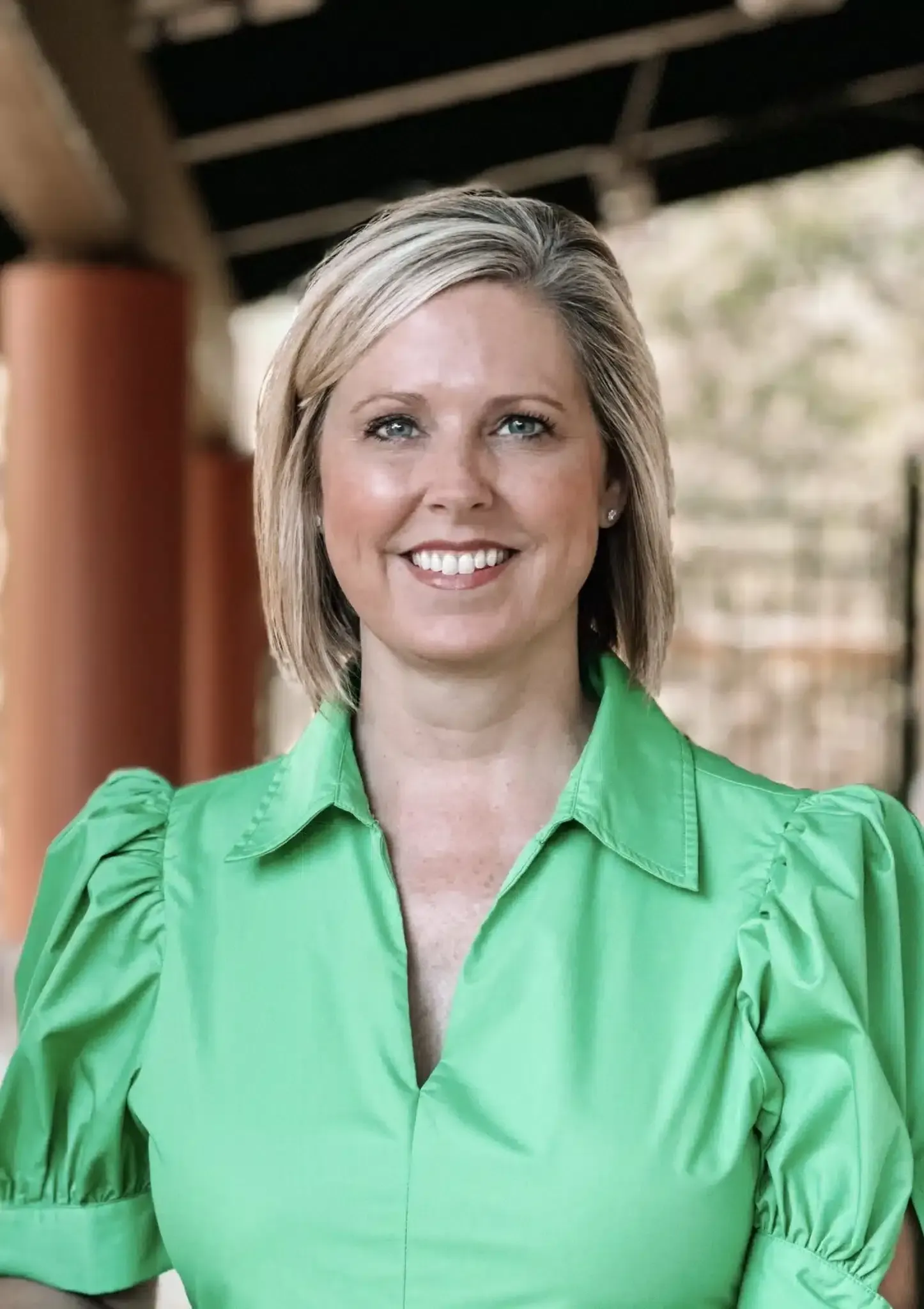 Woman with blonde hair, wearing a green blouse, smiling. Outdoors, background of a building.