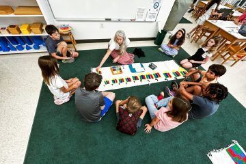 Children and teacher sit in a circle on a carpeted floor, using colorful counting blocks. Classroom setting.