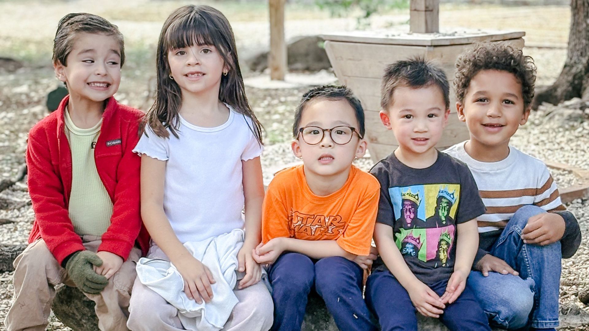 Five children smiling, sitting outside. One wears glasses and an orange shirt.