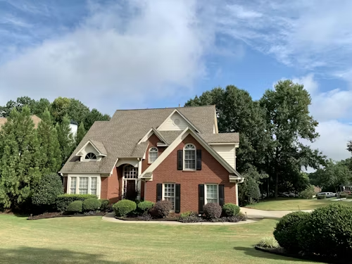 Brick suburban house with beige roof, white-trim windows, and green lawn under a blue sky.