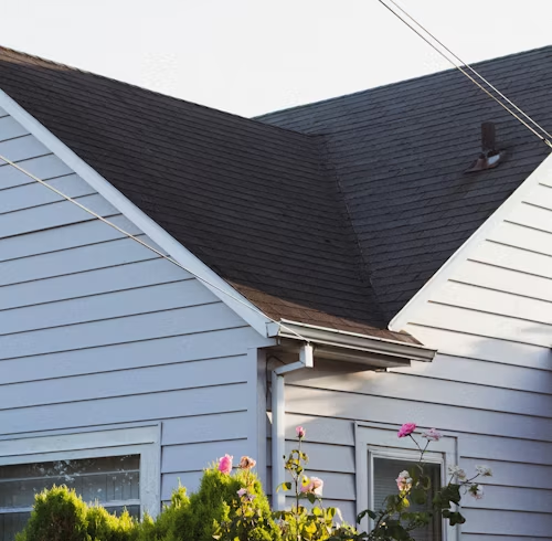 Close-up of a light blue house with a dark roof. A section of the house with white trim, windows, and bushes are visible.