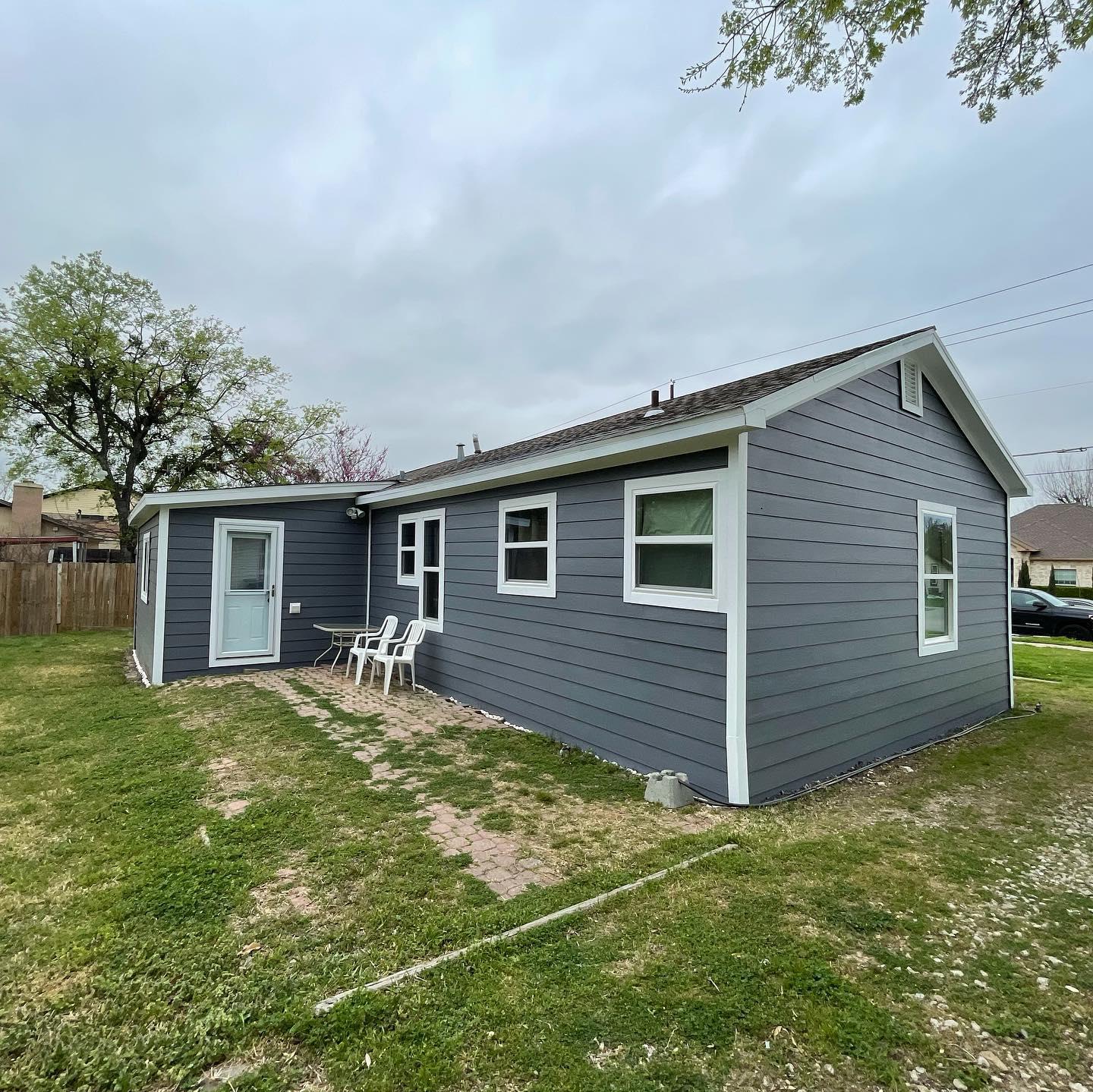 A small gray house with white trim is sitting in the middle of a lush green field.