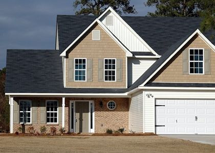Two-story house with dark roof, tan siding, brick accents, and a white garage door against a cloudy sky.