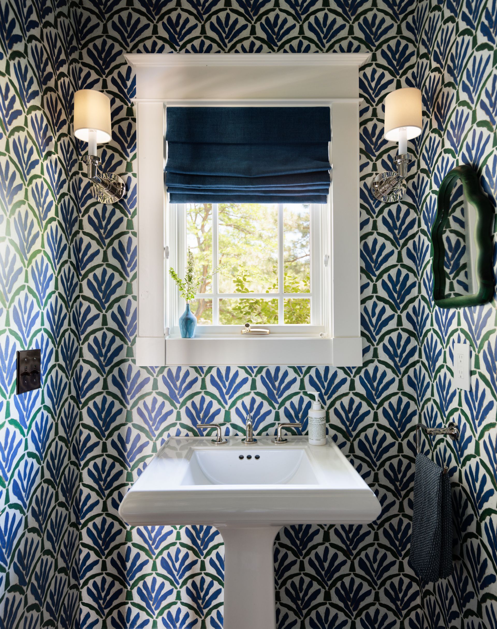 A bathroom with blue and green wallpaper , a sink , a mirror and roman shades of a sink window.