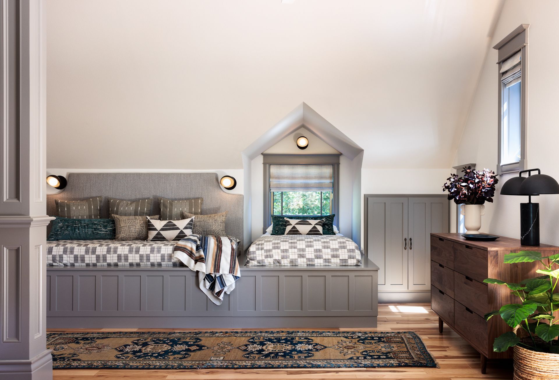 A bedroom with a bed in the corner and a window seat with a dormer window with roman shades