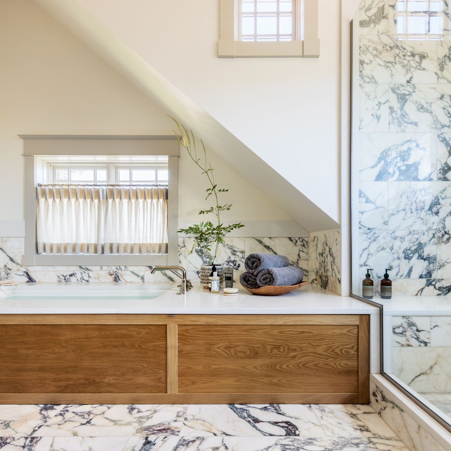 A bathroom with a cafe curtains over the wooden bathtub and marble tiles