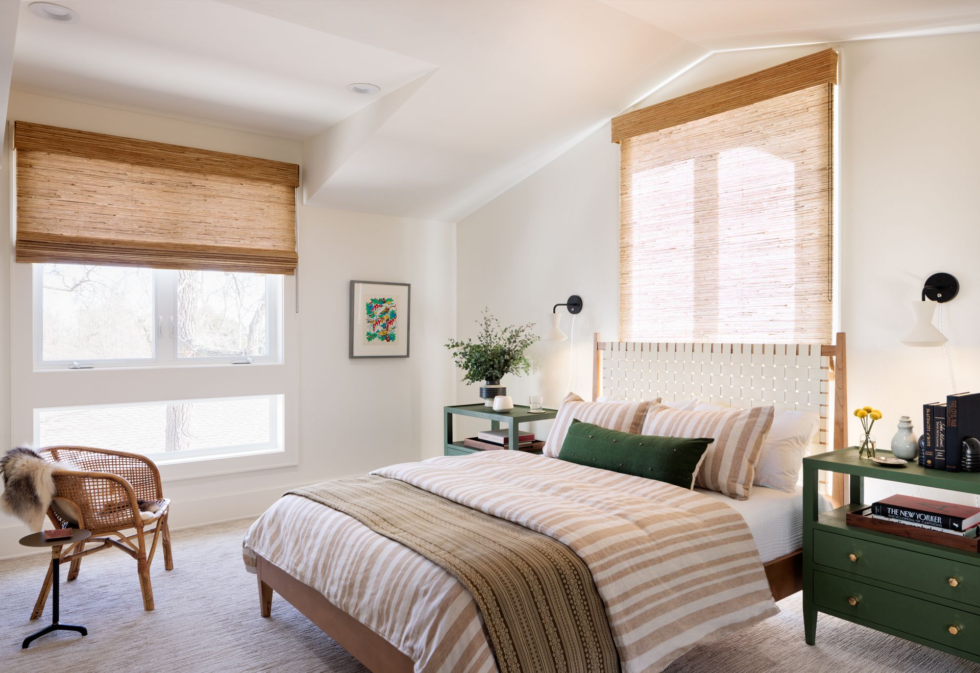 A bedroom with a bed , dresser , chair and two window and woven wood roman shades