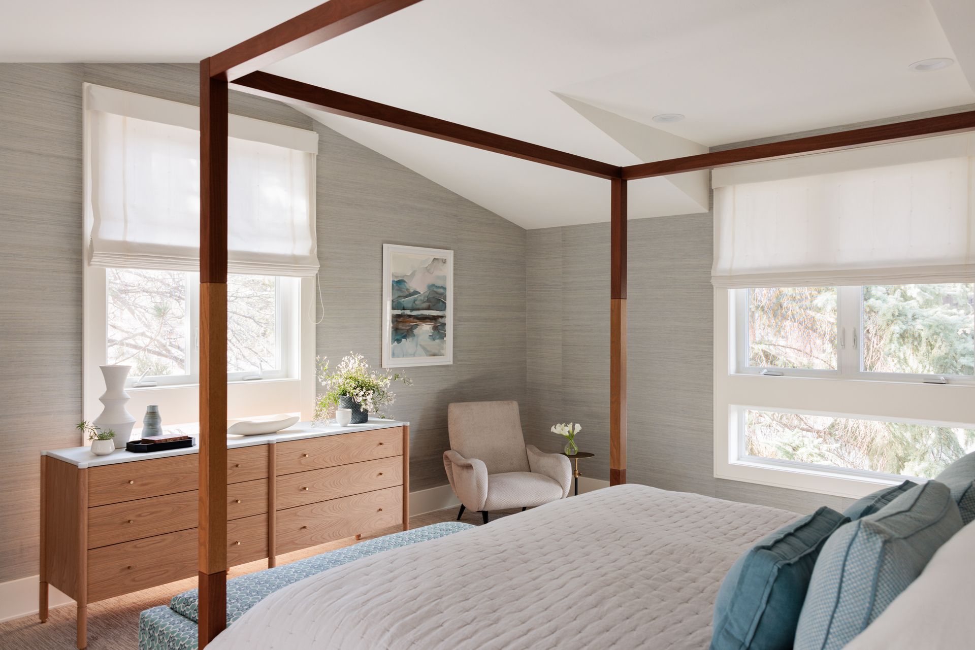 A bedroom with a canopy bed , dresser , chair and windows with white roman shades