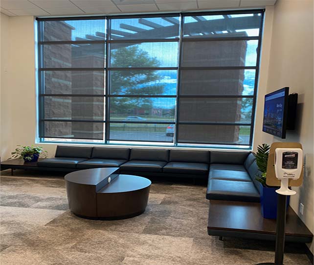 A office lobby with sitting room, roller shades, with a couch , table , and television.