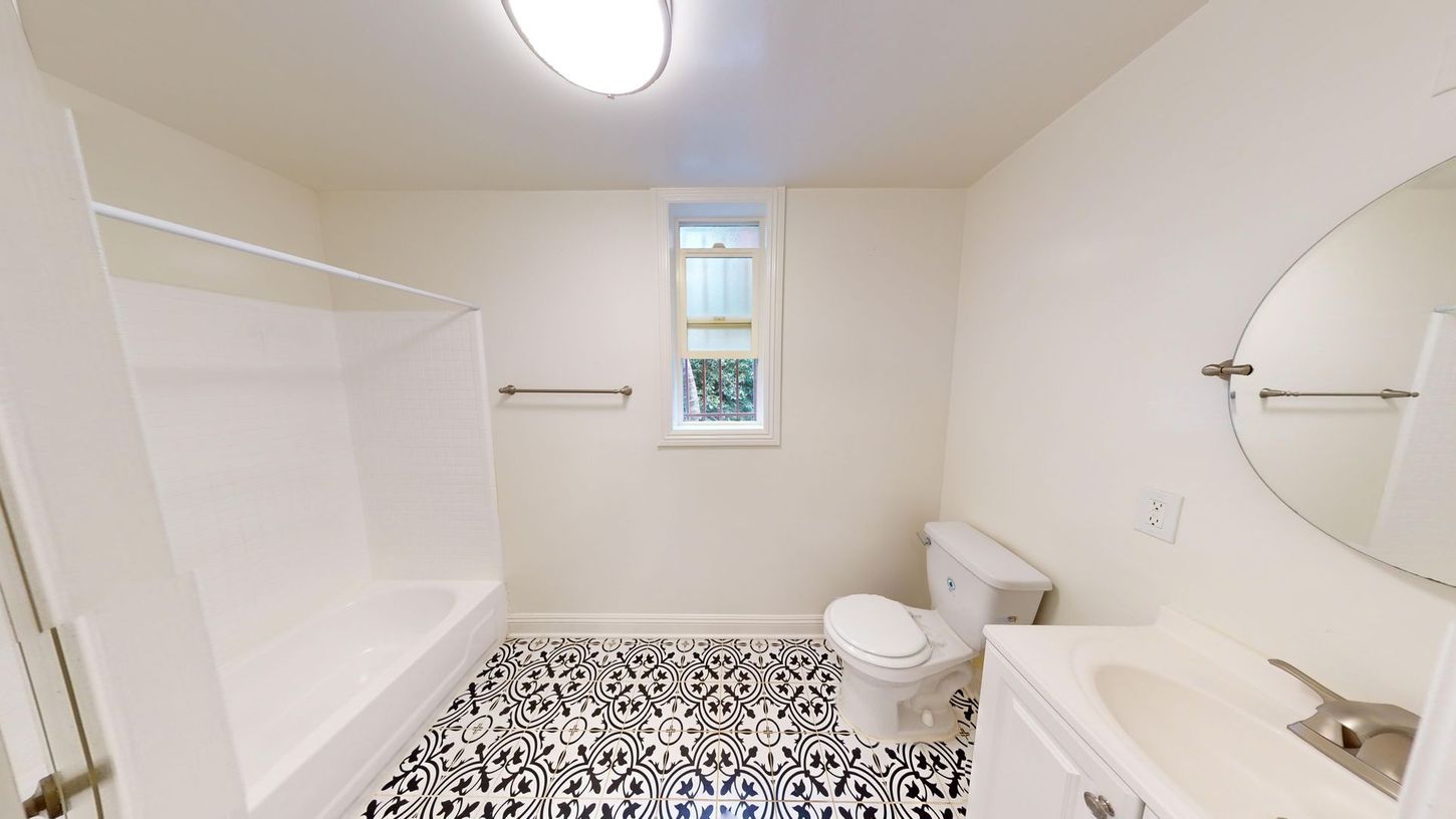 Bathroom with black and white patterned floor, white tub, toilet, and vanity, and a round mirror.