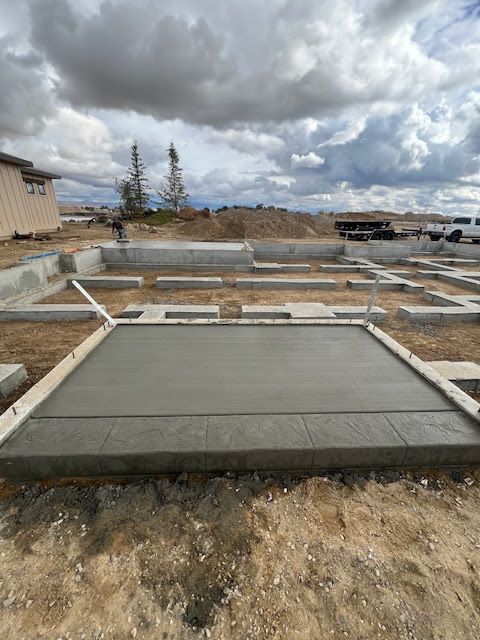A newly poured concrete slab at a residential construction site with dirt, foundations, and a cloudy sky in the background.