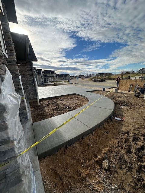 A freshly poured concrete sidewalk curves alongside a new house construction site under a cloudy blue sky.