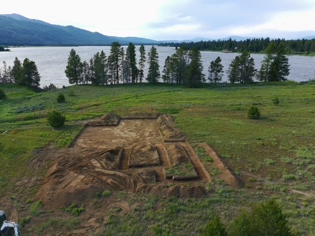 An aerial view of an excavated construction site in a grassy field near a lake and treeline.