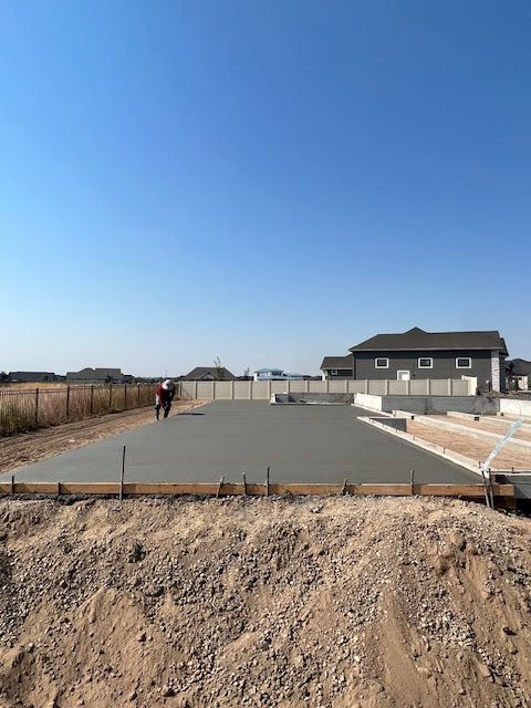 A construction worker levels freshly poured concrete for a home foundation under a clear blue sky.