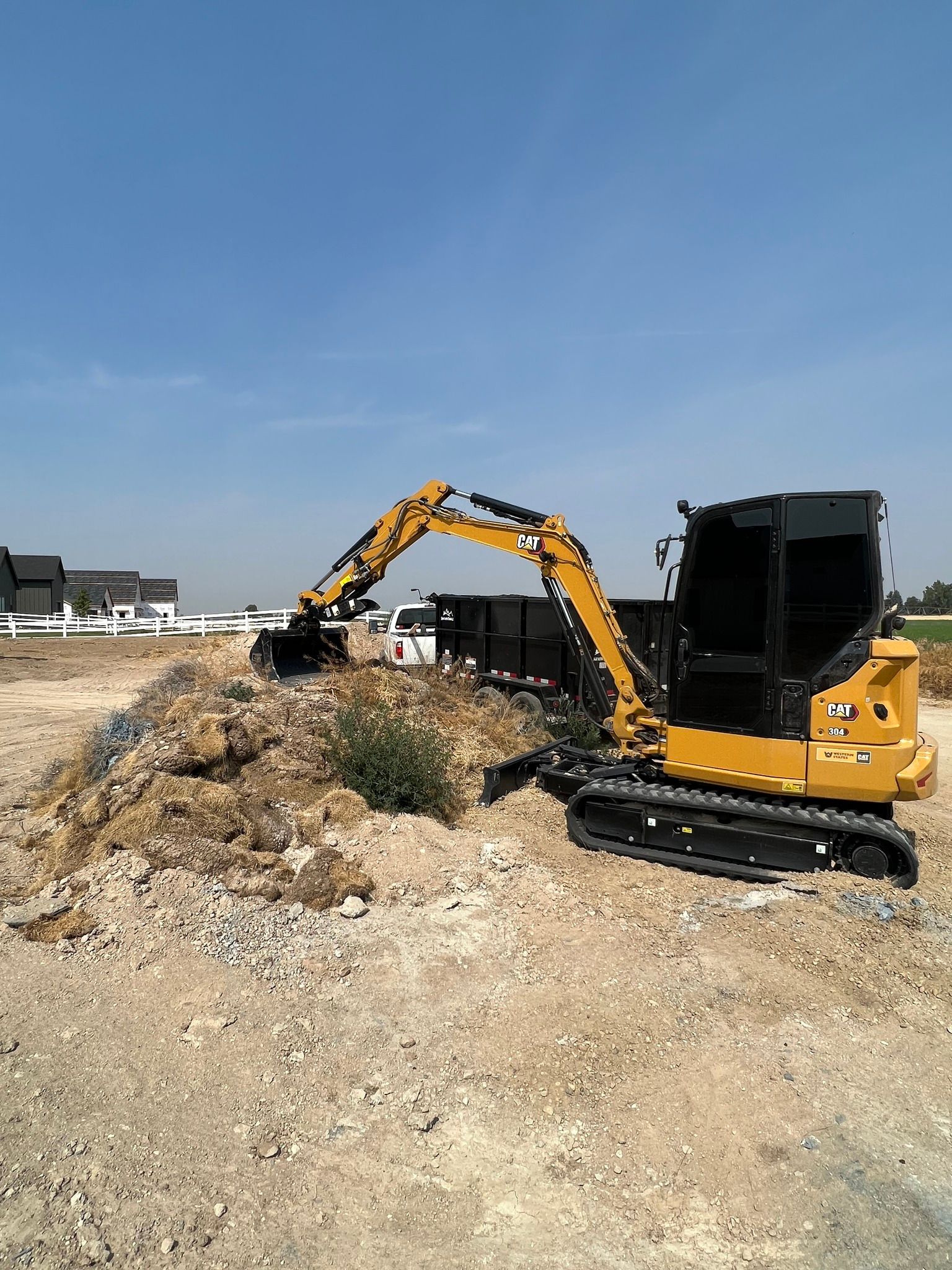 A yellow Caterpillar excavator sits on a dirt construction site, parked next to a large pile of earth and a dump trailer.