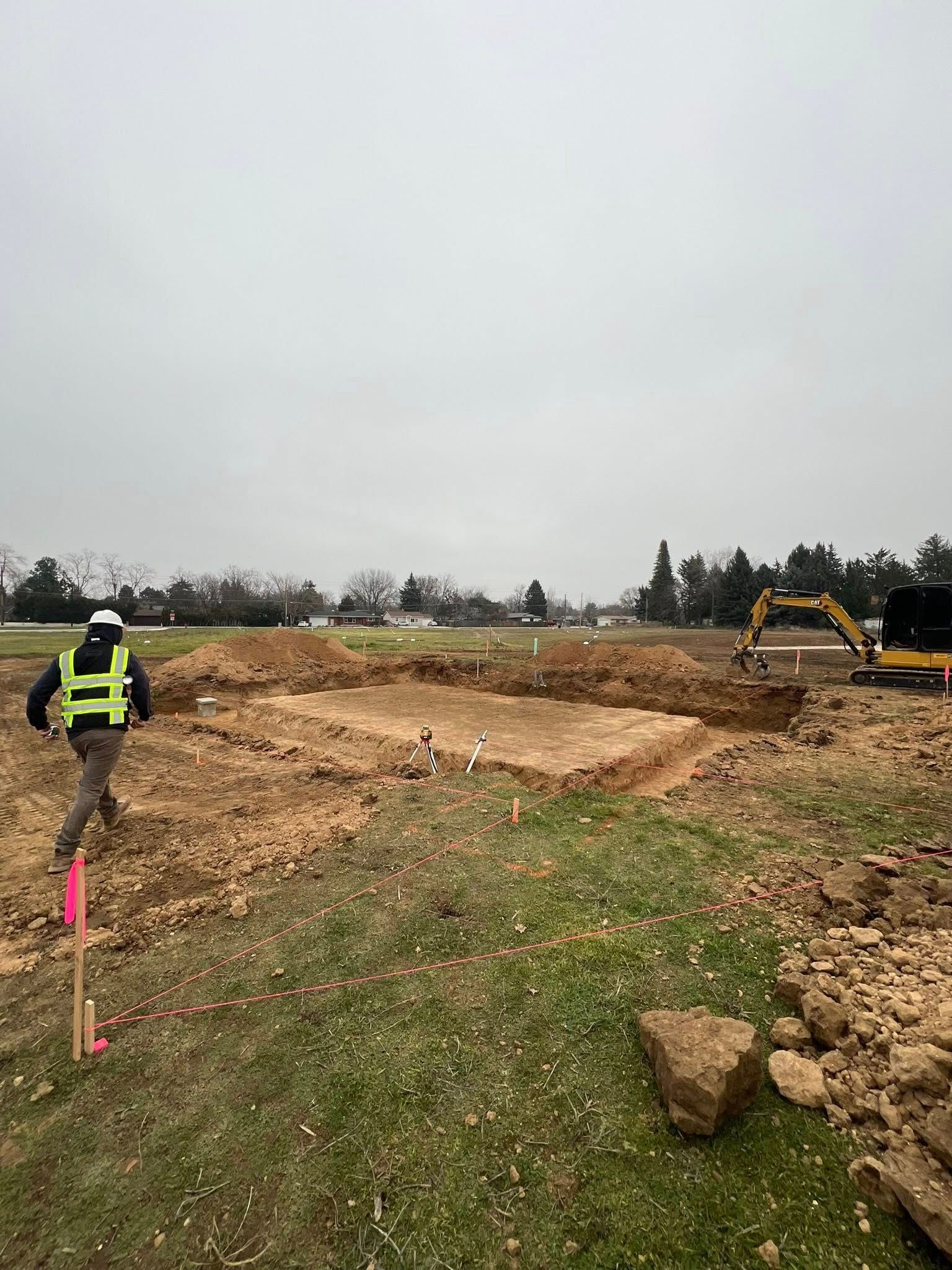 A construction worker in a high-visibility vest walks near a dirt excavation site with yellow equipment in the distance.