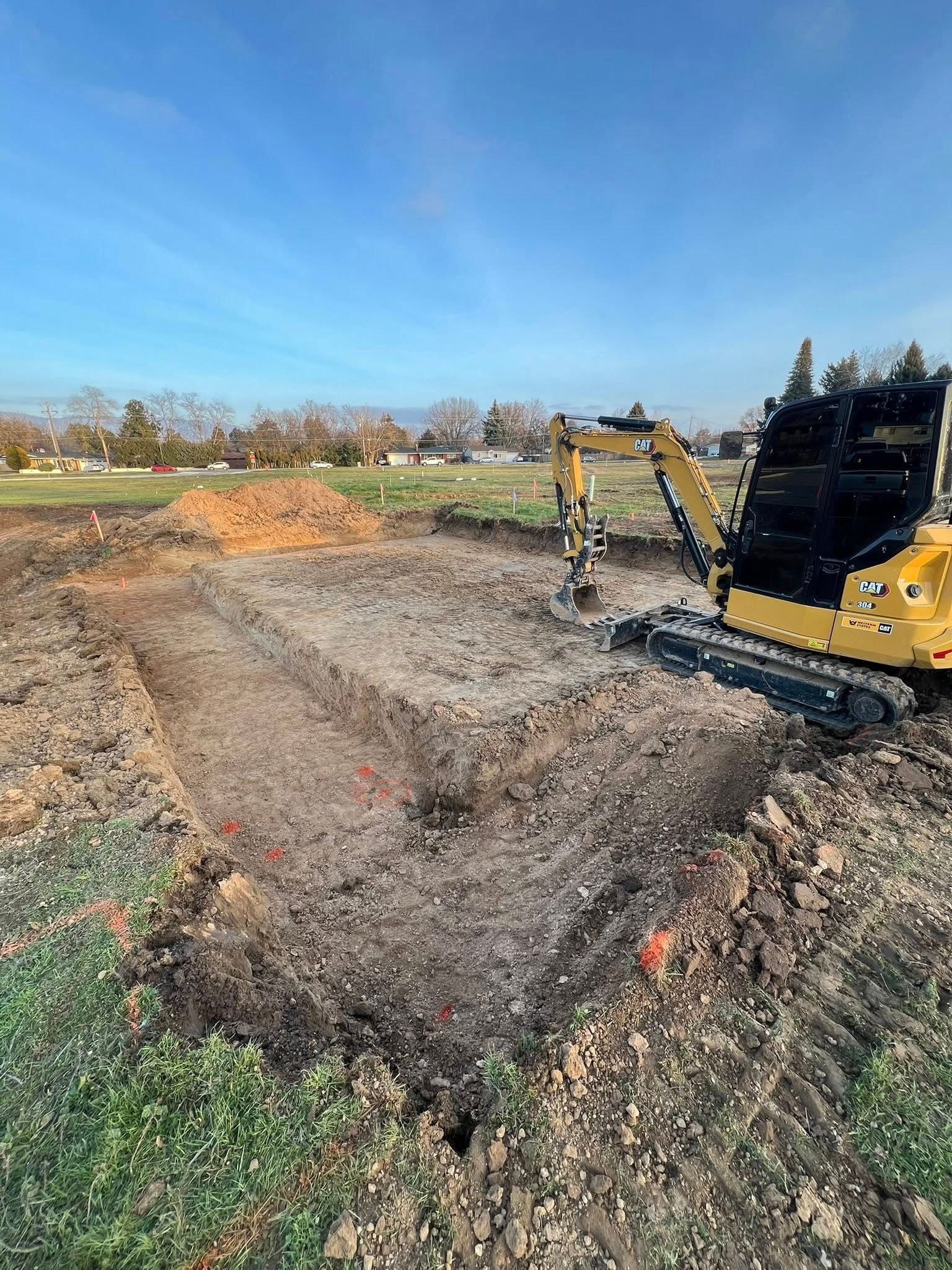 A yellow excavator sits in a freshly dug rectangular foundation pit on a sunny, clear day.