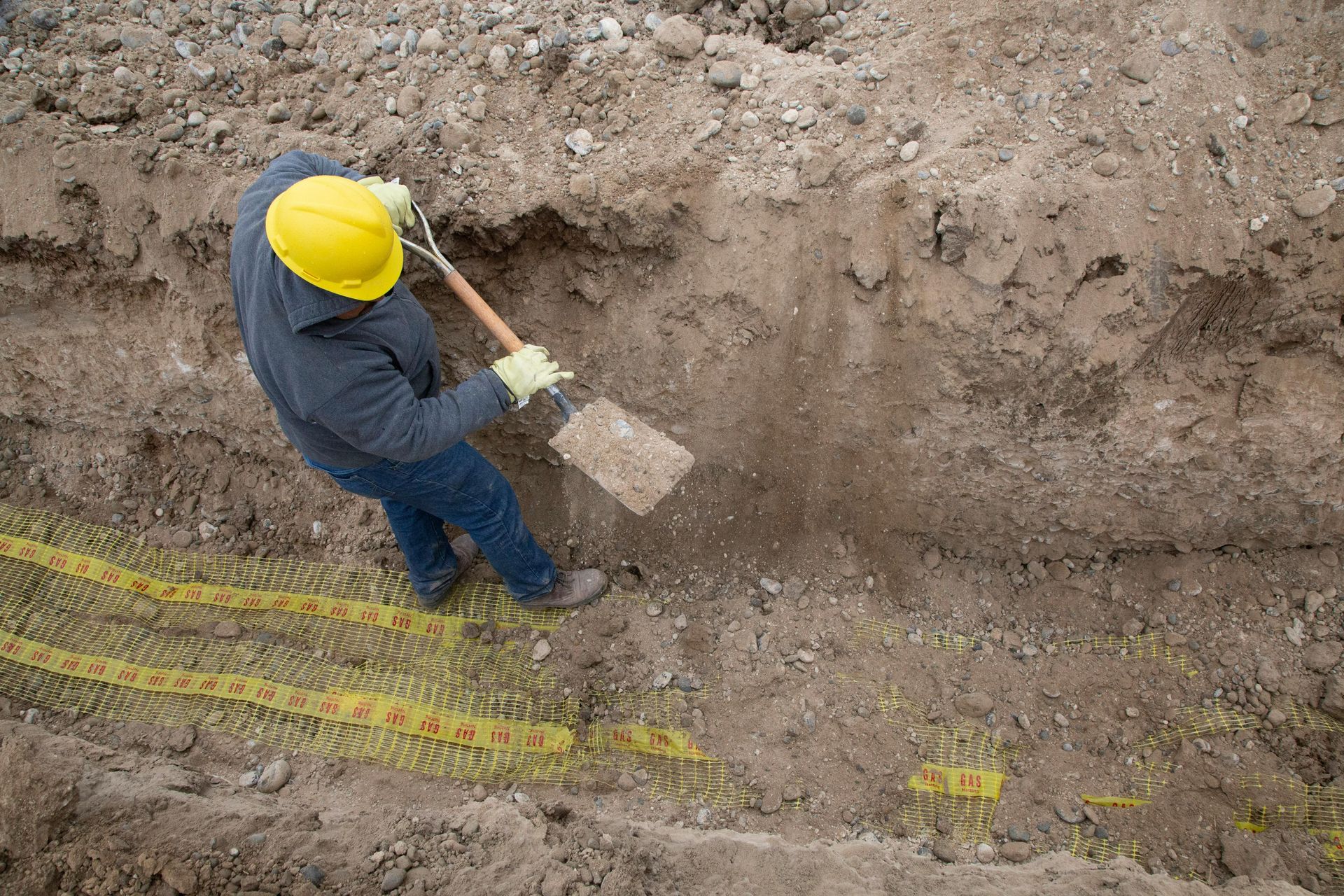 A person in a yellow hard hat and work clothes uses a shovel to clear dirt inside a deep trench with yellow safety mesh.