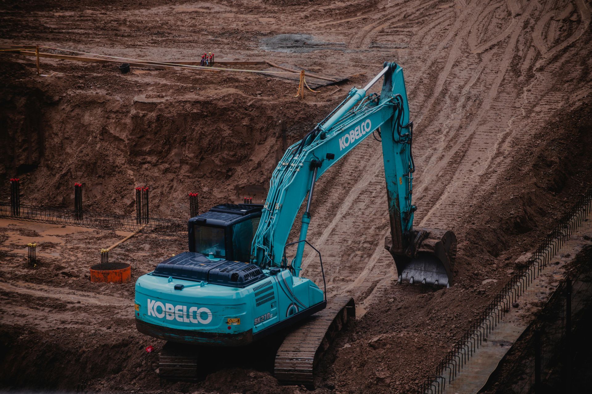 A blue Kobelco excavator sits on dirt at a construction site, with its bucket resting on a sloped embankment.