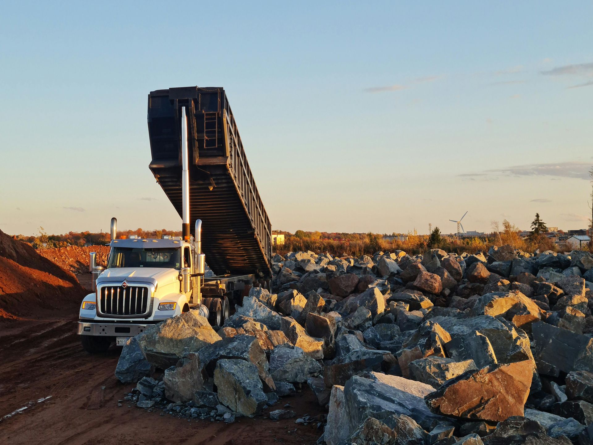 A white dump truck with its bed fully raised stands in a field of large rocks and red soil under a clear sky.