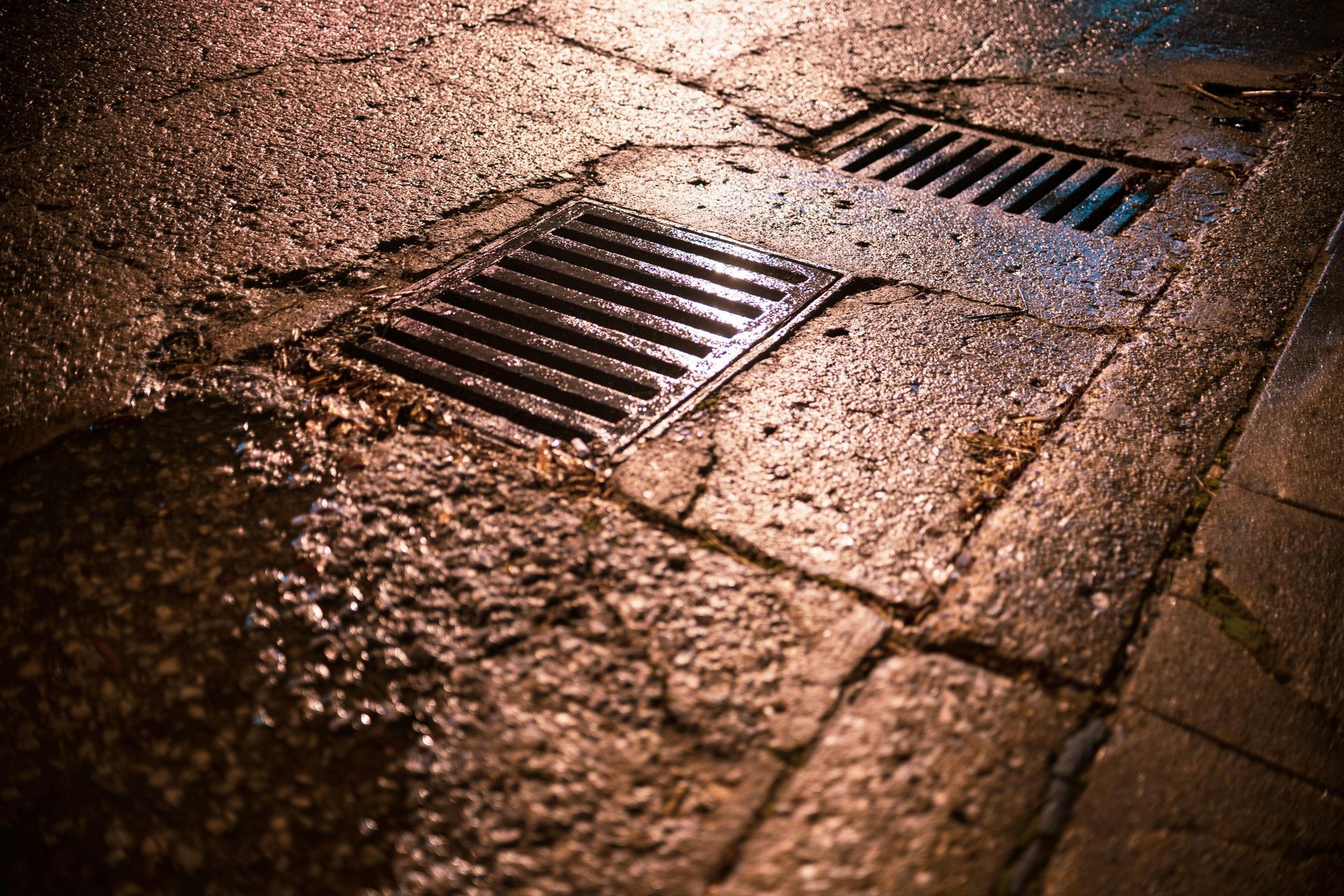 A metal storm drain grate set into a wet, textured stone or asphalt surface at night under warm artificial lighting.