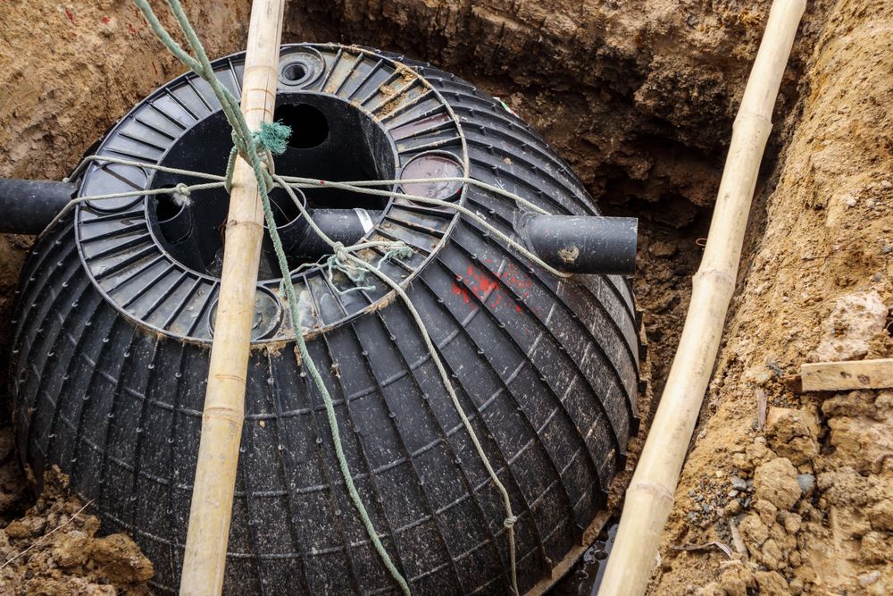 A black, domed plastic septic tank being installed in a dirt trench, secured by bamboo poles and rope.