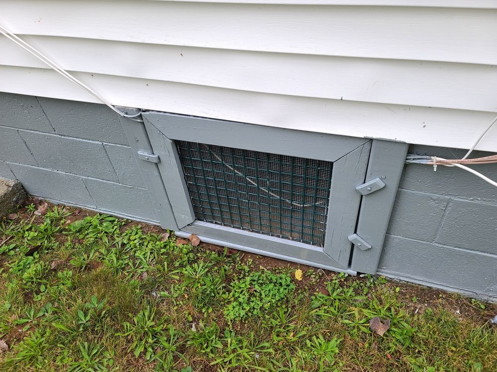 A rectangular grey-painted foundation vent with wire mesh installed in a concrete block wall beneath white siding.