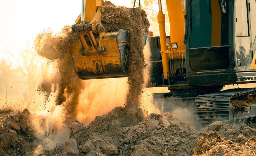 A yellow excavator's bucket dumps dirt onto a pile at a construction site, creating a cloud of dust.