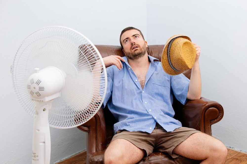 A man is sitting in a chair holding a hat in front of a fan.